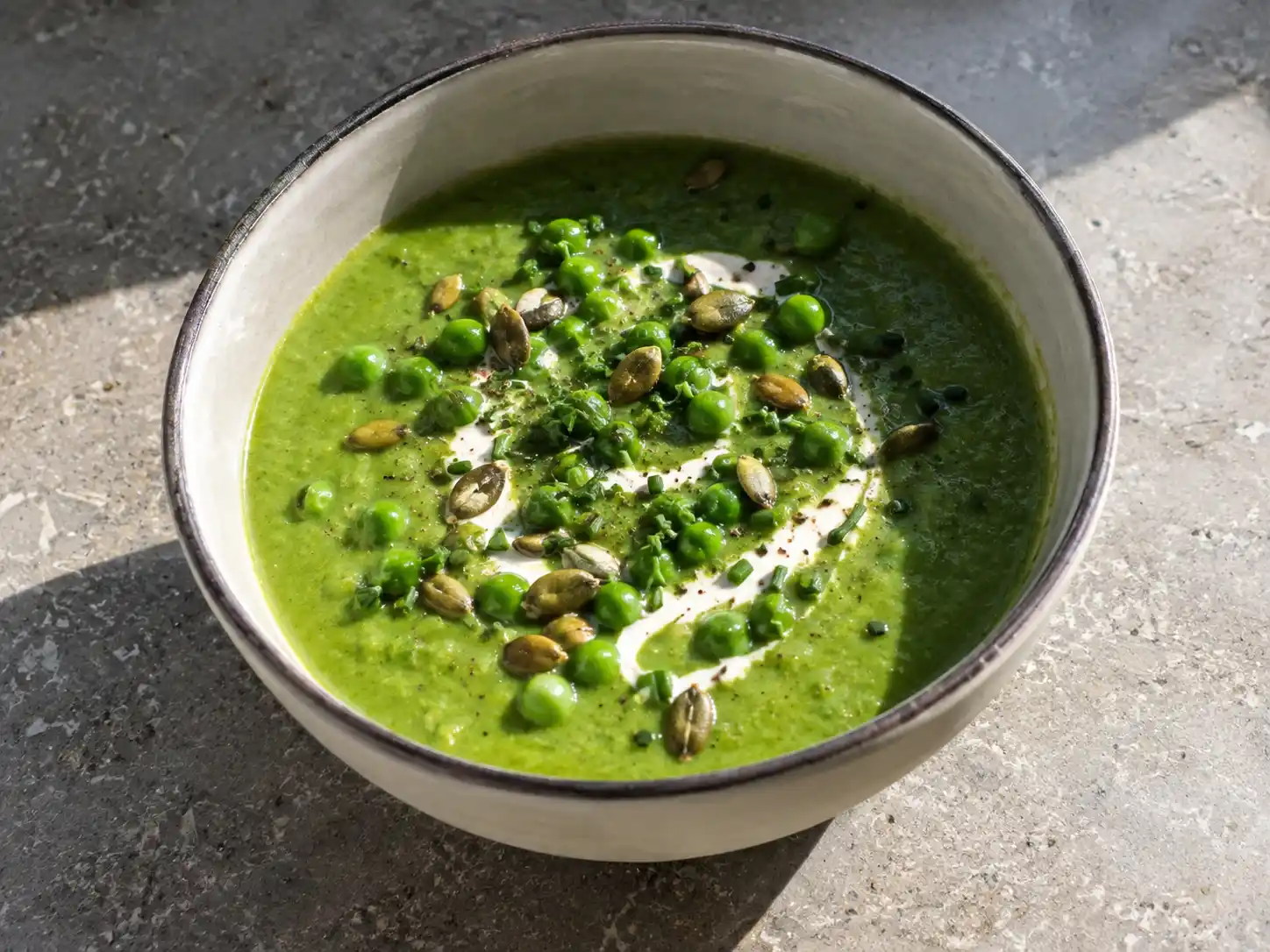 Grön ärtsoppa i djup skål med crème fraiche, gröna ärtor, pumpakärnor och hackad gräslök ovanpå