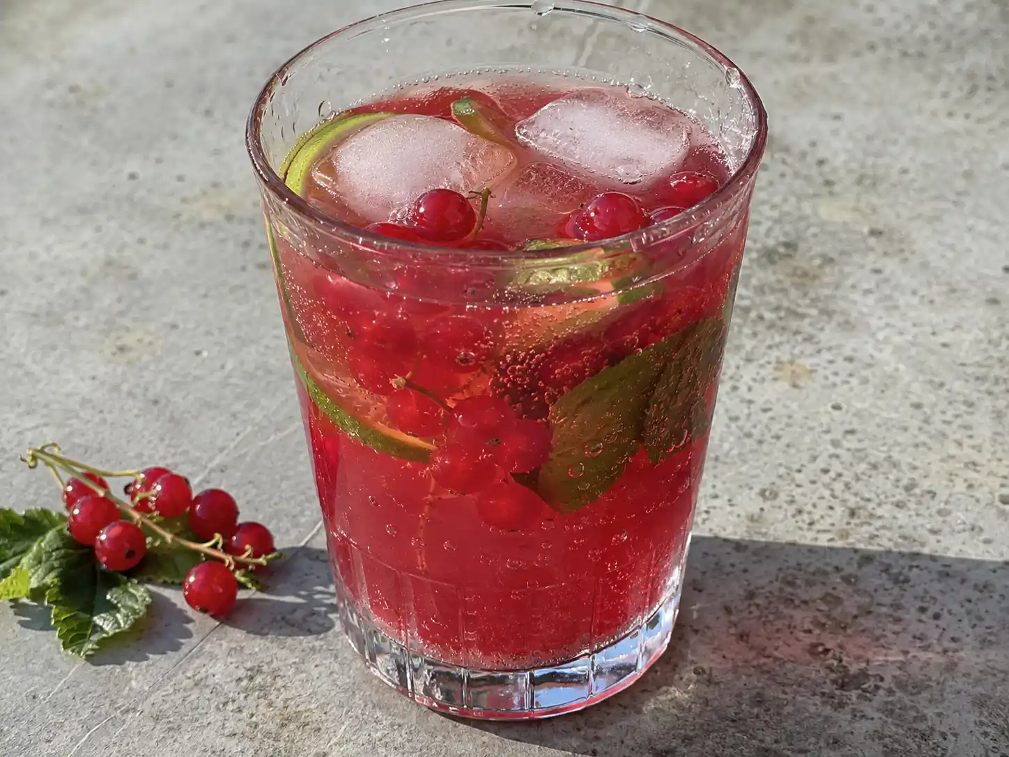 röda vinbär drink i glas med lime, mynta, is och röda vinbärsklasar på enkelt bord
